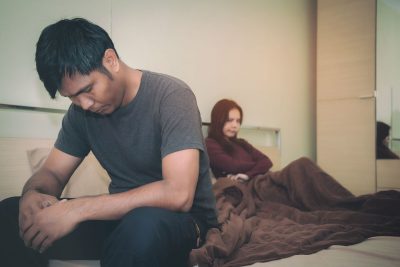 Fire fighter out of uniform sitting away from his spouse on the bed with an upset look